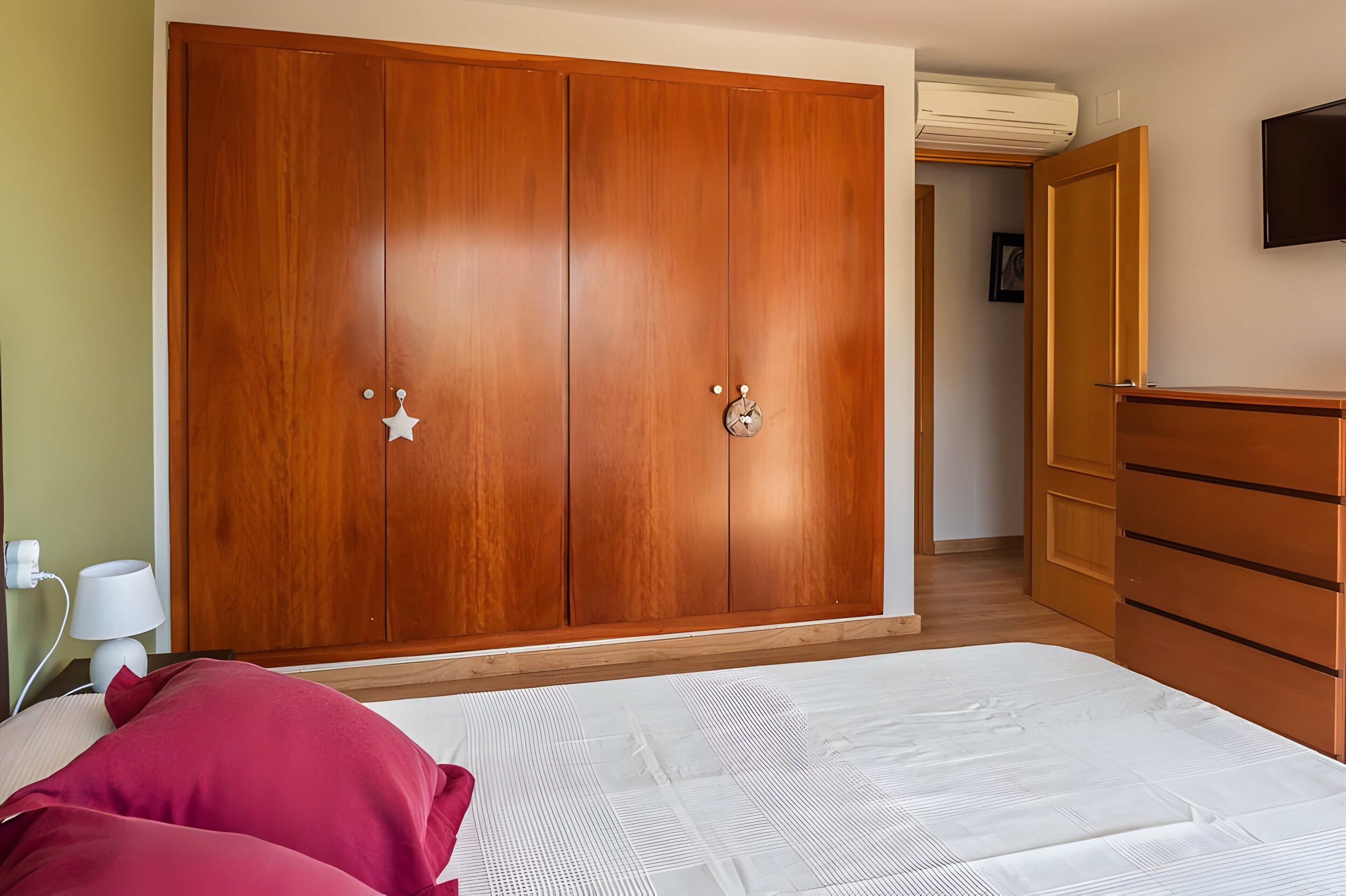 A bedroom with a wooden wardrobe, a bed with white bedding, a dresser, and a television on the wall.