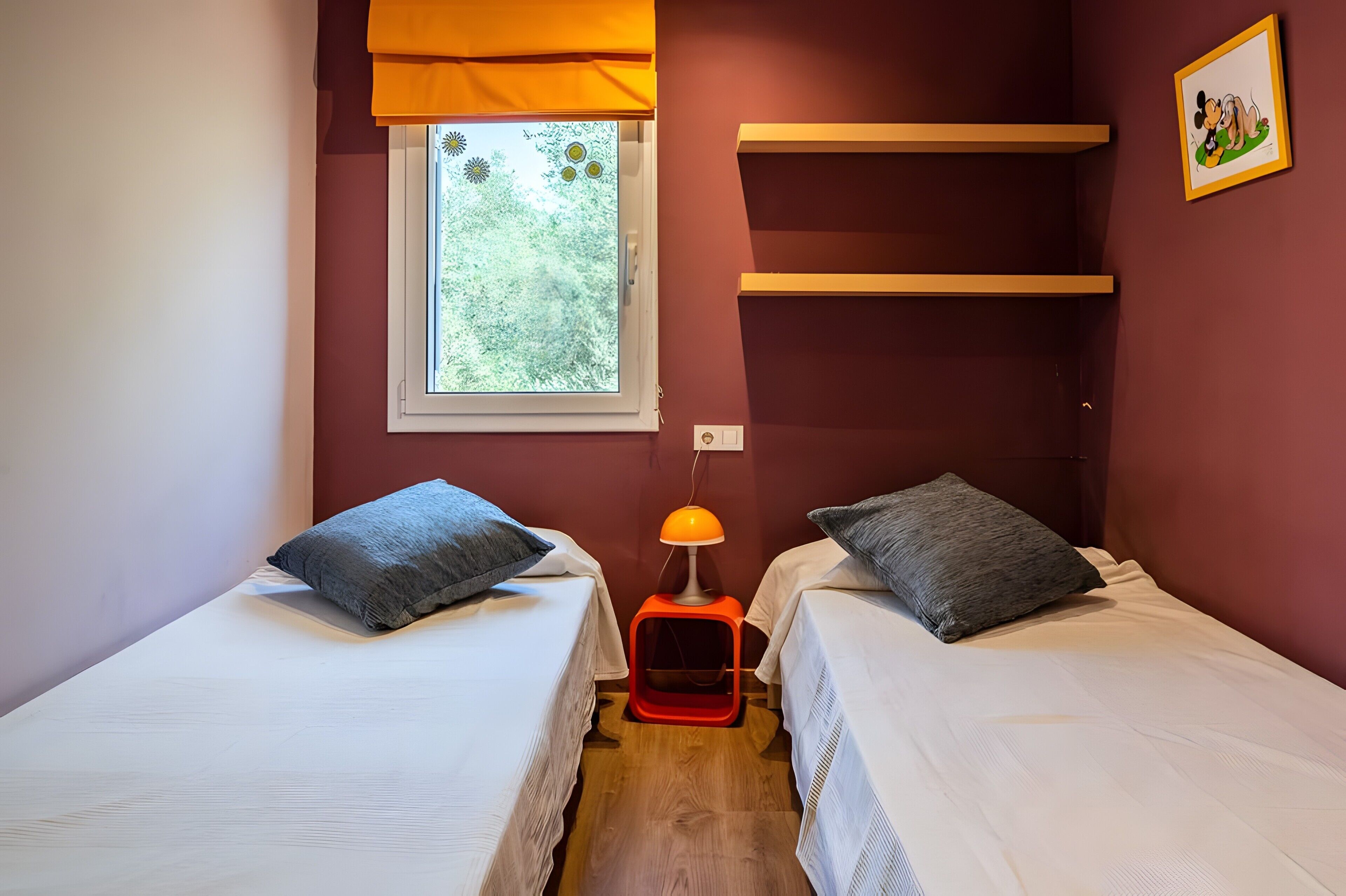 A room with two beds, a window with a view of greenery, a wall-mounted shelf, and a picture.