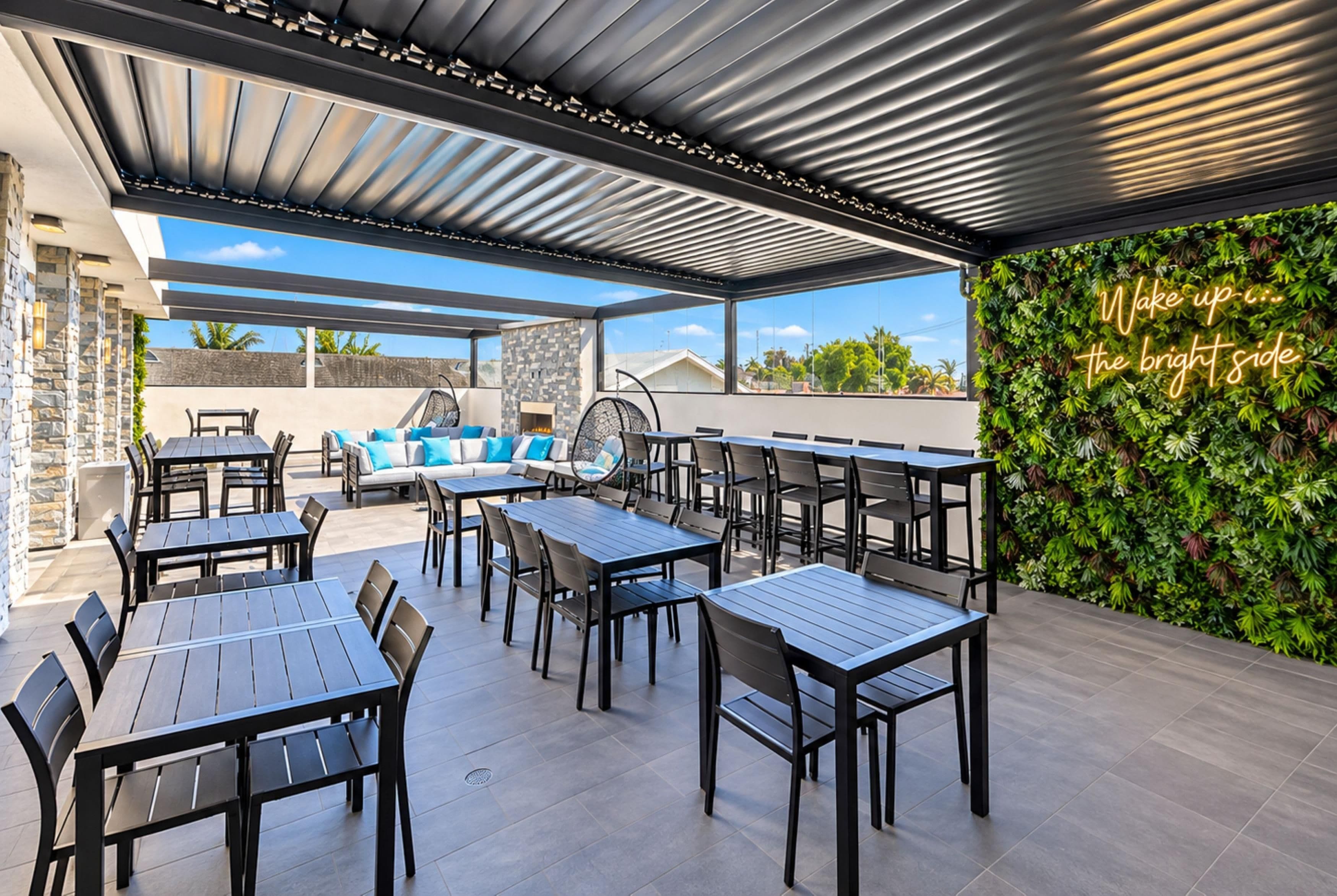 An outdoor patio area with tables and chairs, a seating area with cushions, a stone wall, and a wall with a neon sign.