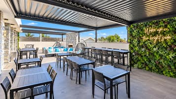 An outdoor patio area with tables and chairs, a seating area with cushions, a stone wall, and a wall with a neon sign.