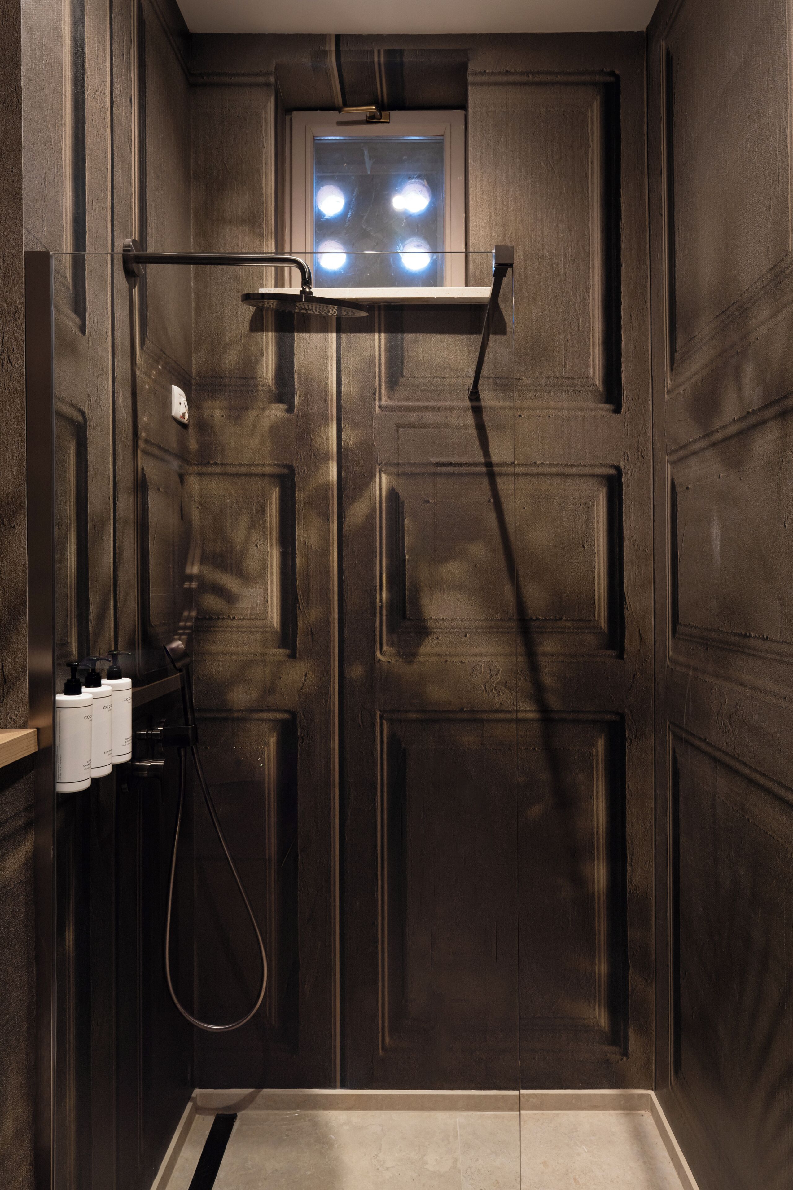 Un baño con diseño de paneles de madera oscura, zona de ducha y ventana con luces.