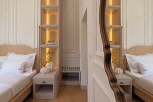 Quarto de hotel com duas camas, mesas de cabeceira e uma estante central com livros e objetos decorativos.