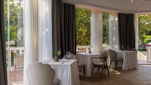 A restaurant with multiple tables set for dining, large windows with curtains, and a view of greenery outside. - Heritage Hotel Zagreb Dubrovik (Dubrovnik)