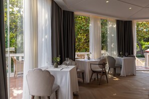 A restaurant with multiple tables set for dining, large windows with curtains, and a view of greenery outside. - Heritage Hotel Zagreb Dubrovik (Dubrovnik)