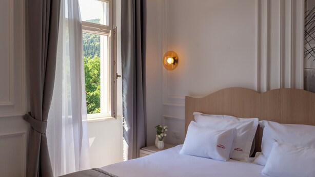 A hotel room with a bed, a nightstand, a vase with flowers, a window with curtains, and a wall-mounted light fixture. - Heritage Hotel Zagreb Dubrovik (Dubrovnik)