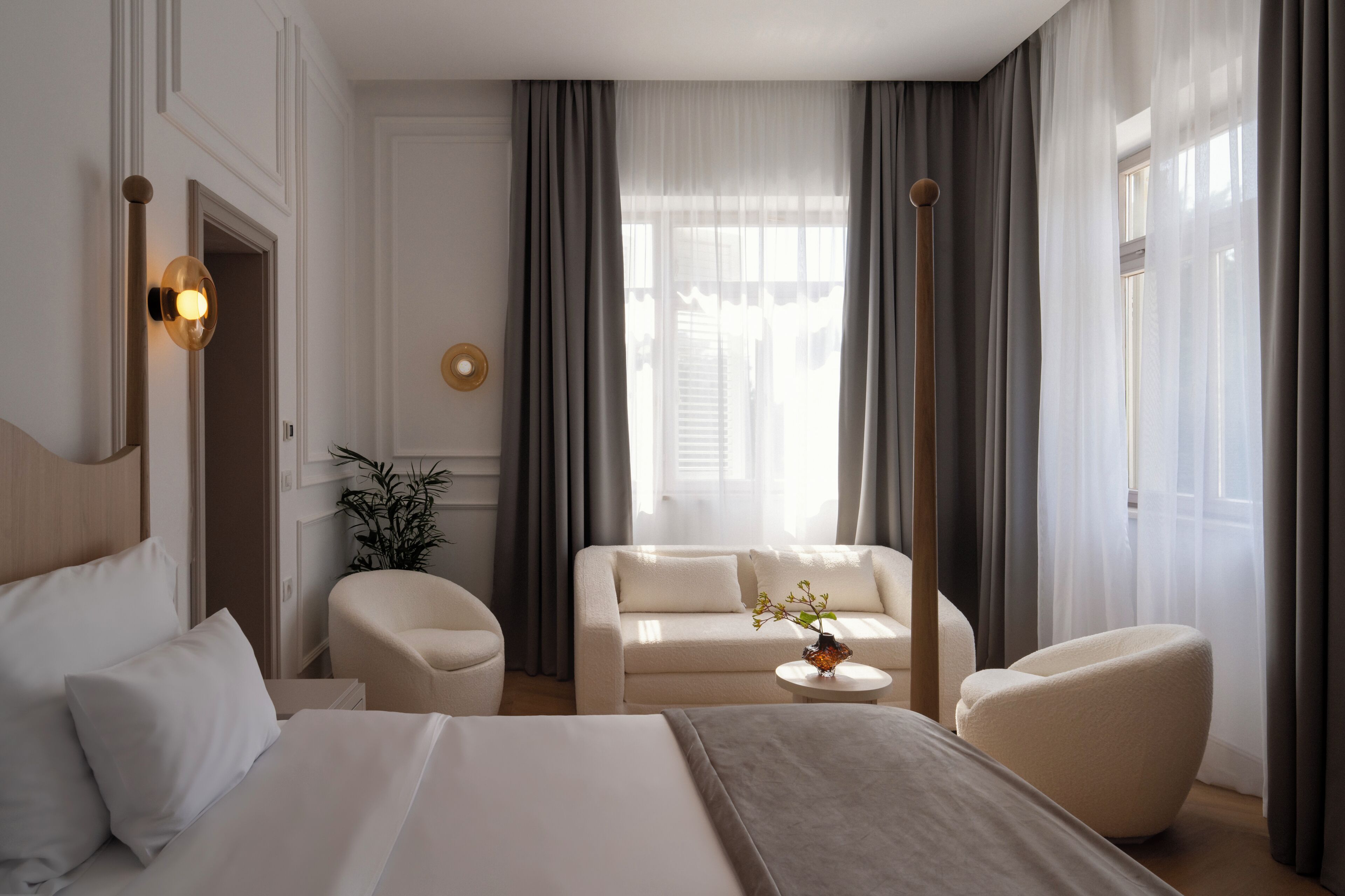 A hotel room with a bed, two armchairs, a sofa, a small table with a vase, and a window with curtains.