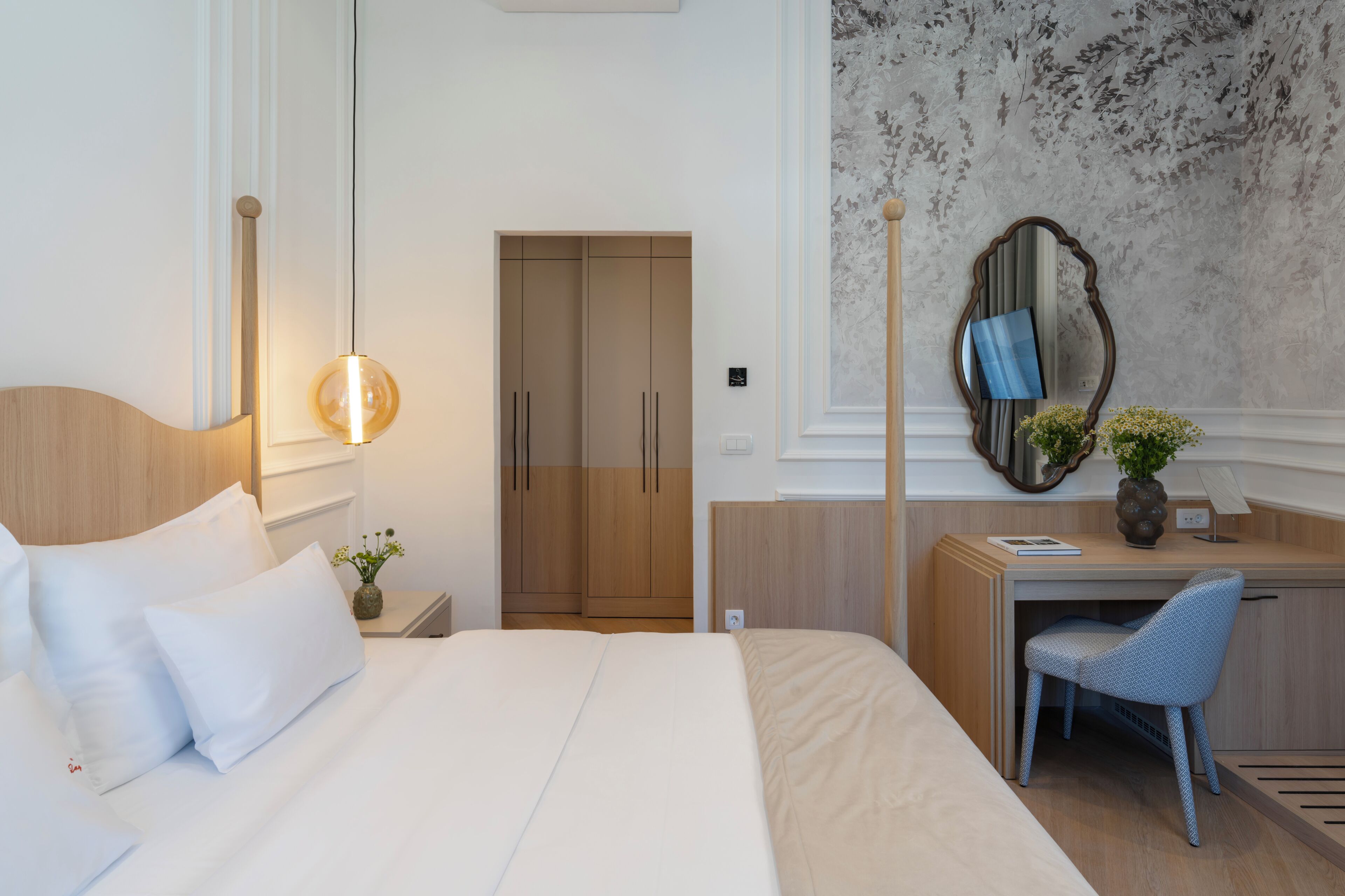 A hotel room with a bed, a desk, a chair, a mirror, and a wall with a floral pattern.