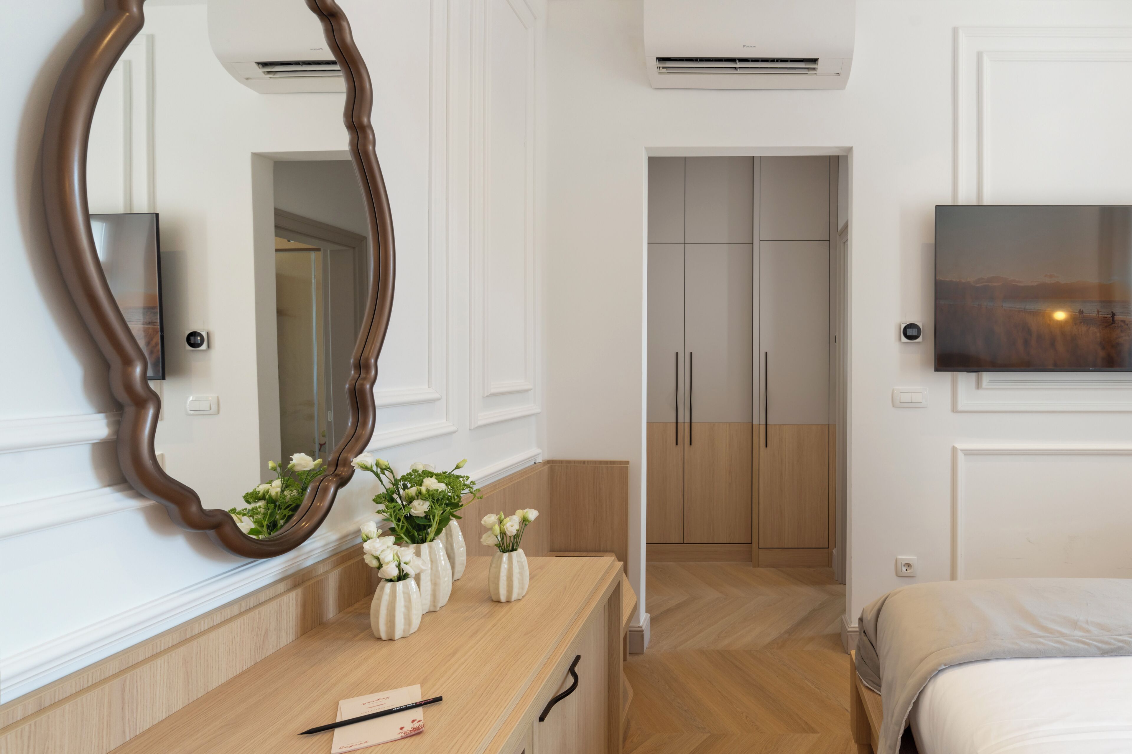 A modern bedroom with a wooden dresser, a large mirror, and a flat-screen TV mounted on the wall.