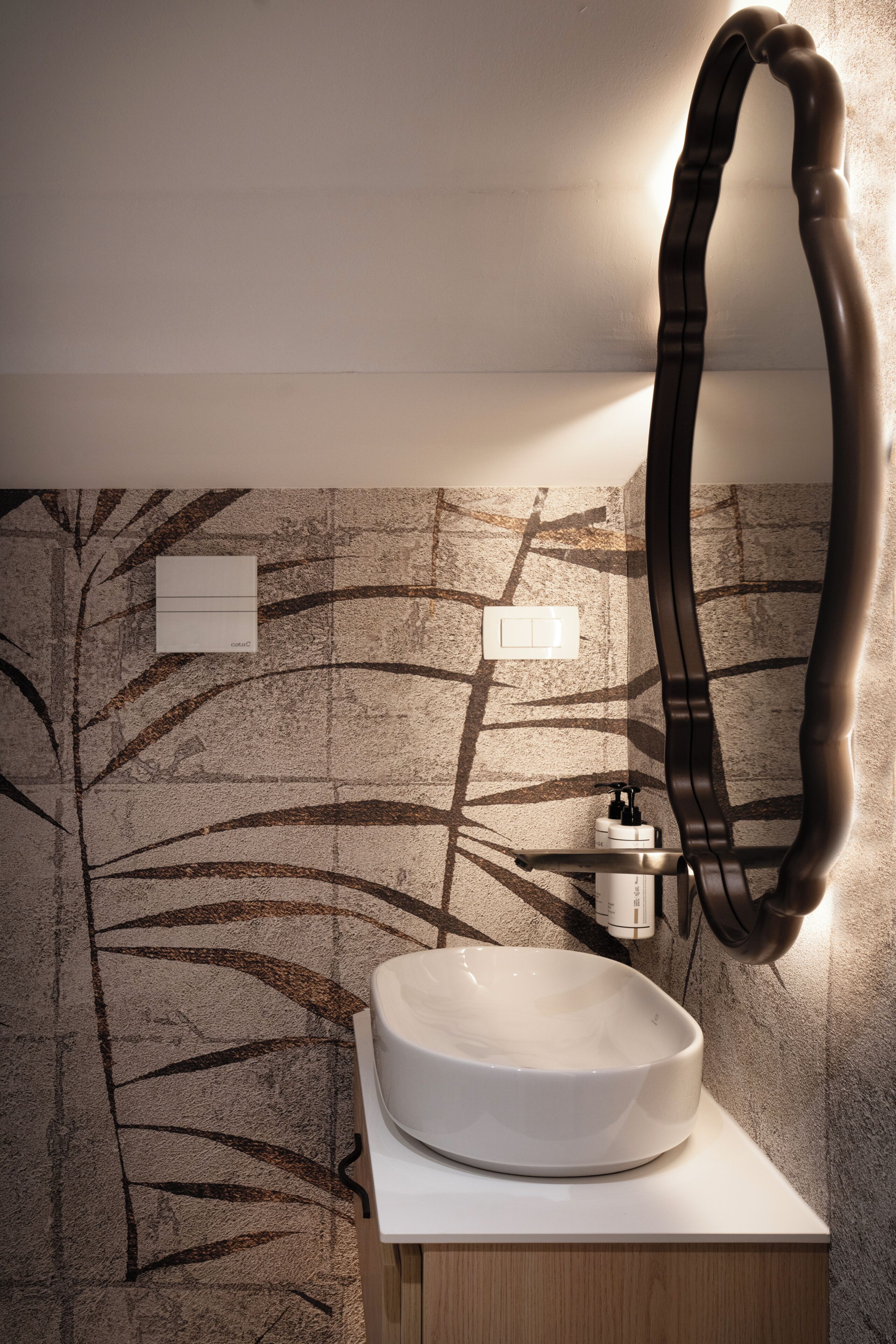Un baño de diseño moderno, con un espejo grande, un lavamanos blanco y una pared con diseño de hojas.