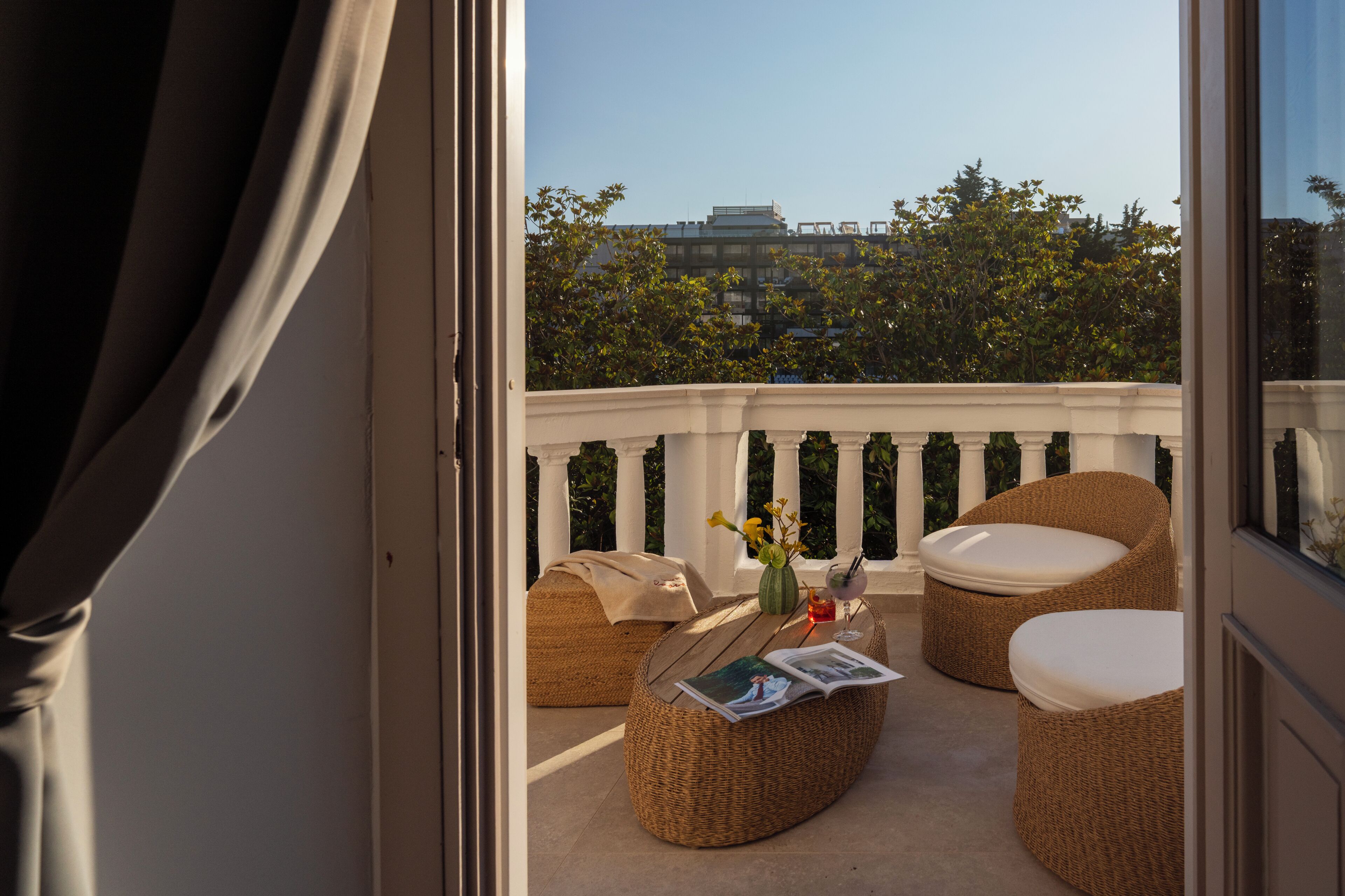 Un balcón con mobiliario de mimbre, una mesa pequeña con una bebida y vistas a edificios y árboles.