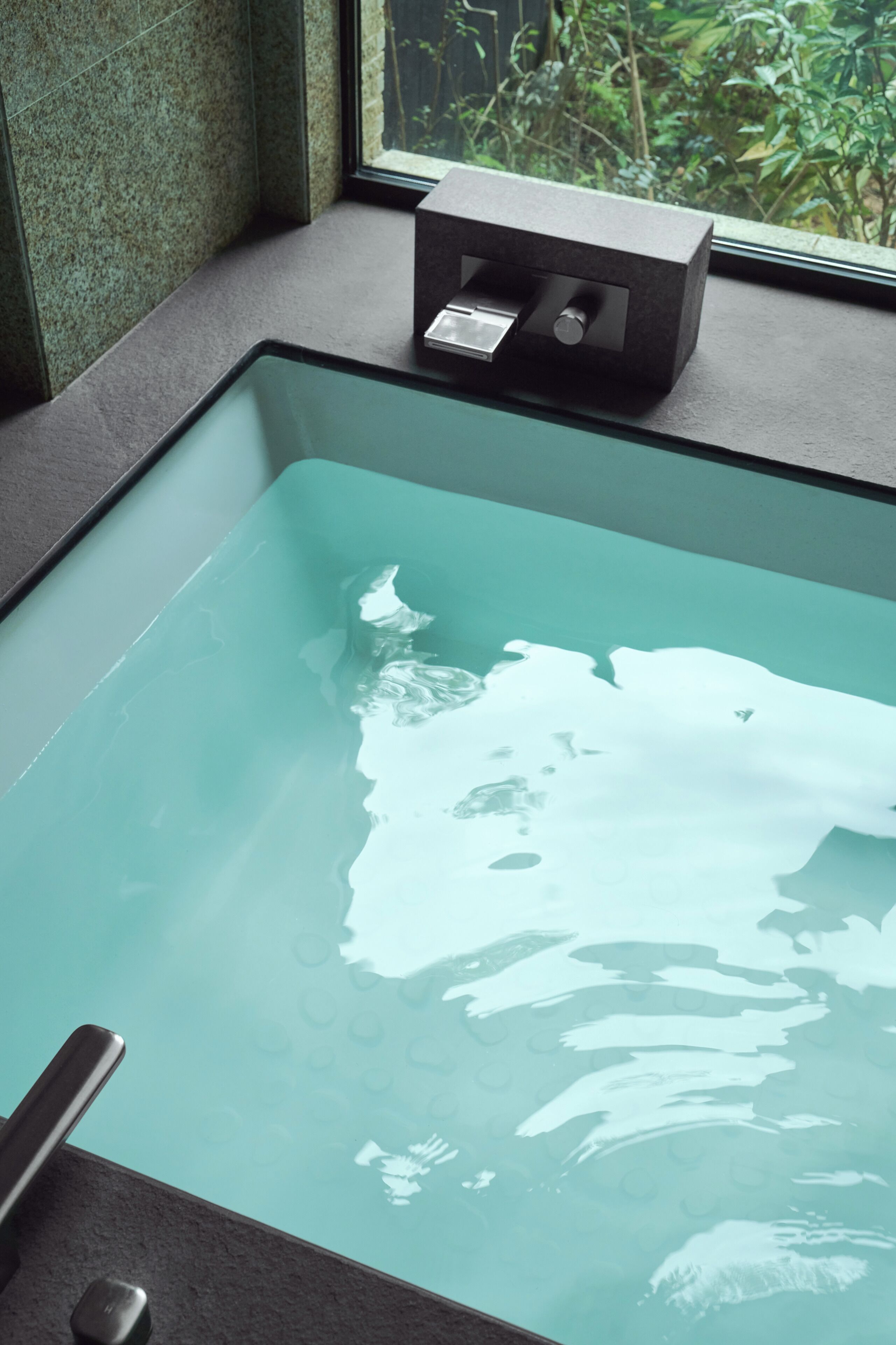 A rectangular glass bathtub with a faucet, positioned near a window with a view of greenery outside.