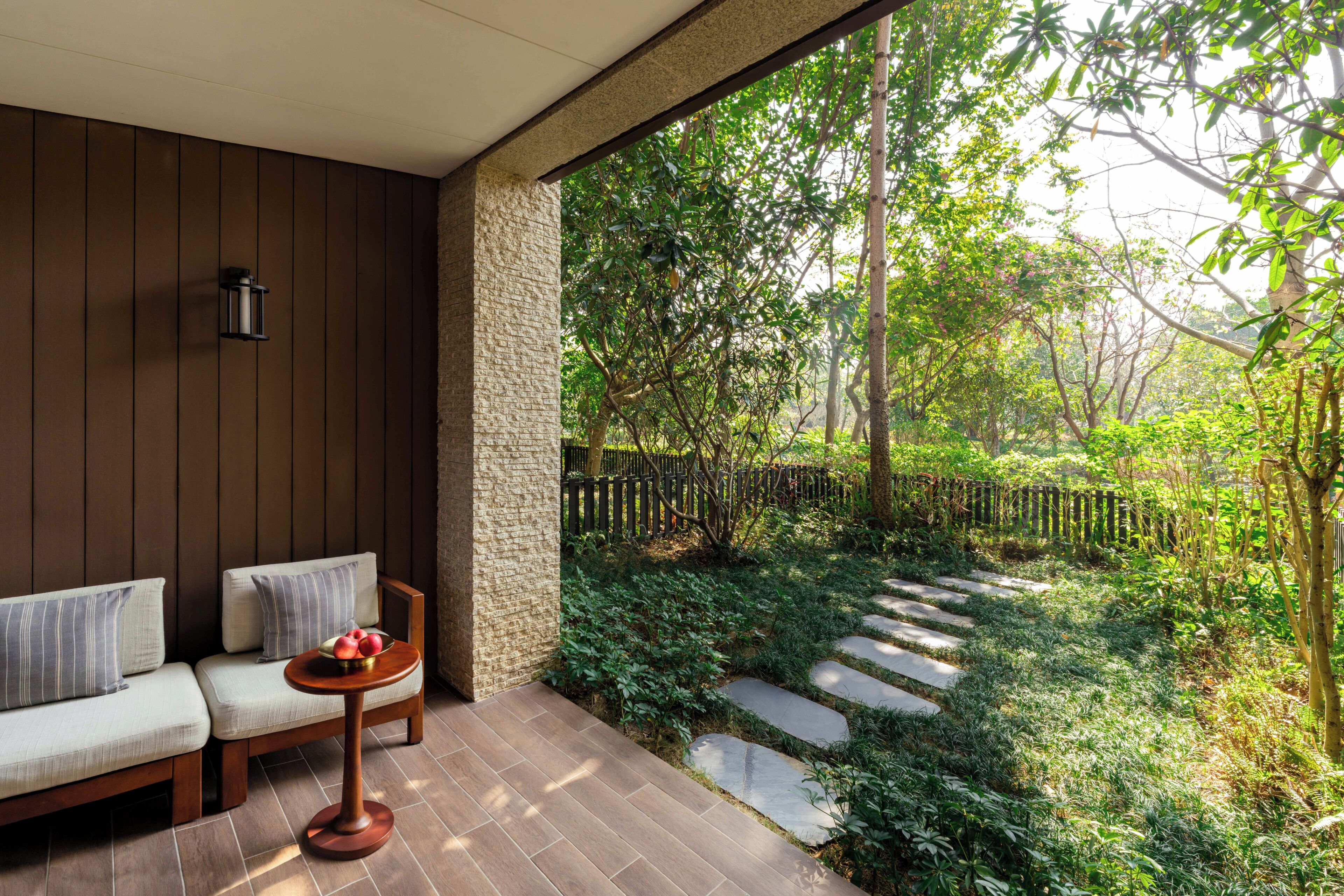 A covered patio with a sofa, a small table with fruit, and a stone pathway leading to a garden.