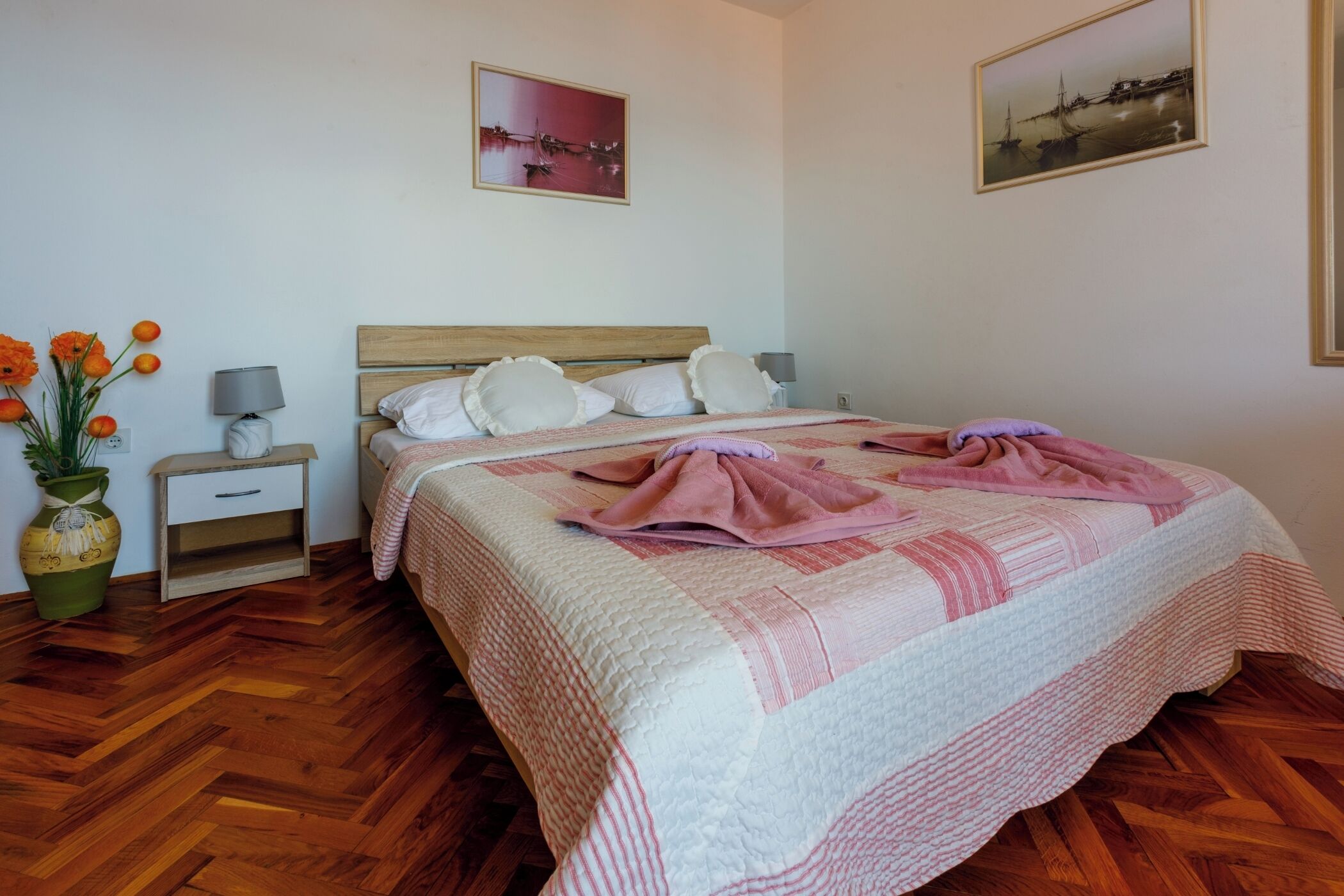 Une chambre à coucher bien rangée, avec un plancher en bois, un lit recouvert d’une courtepointe, une table de chevet avec une lampe, un vase rempli de fleurs et deux tableaux encadrés accrochés au mur.