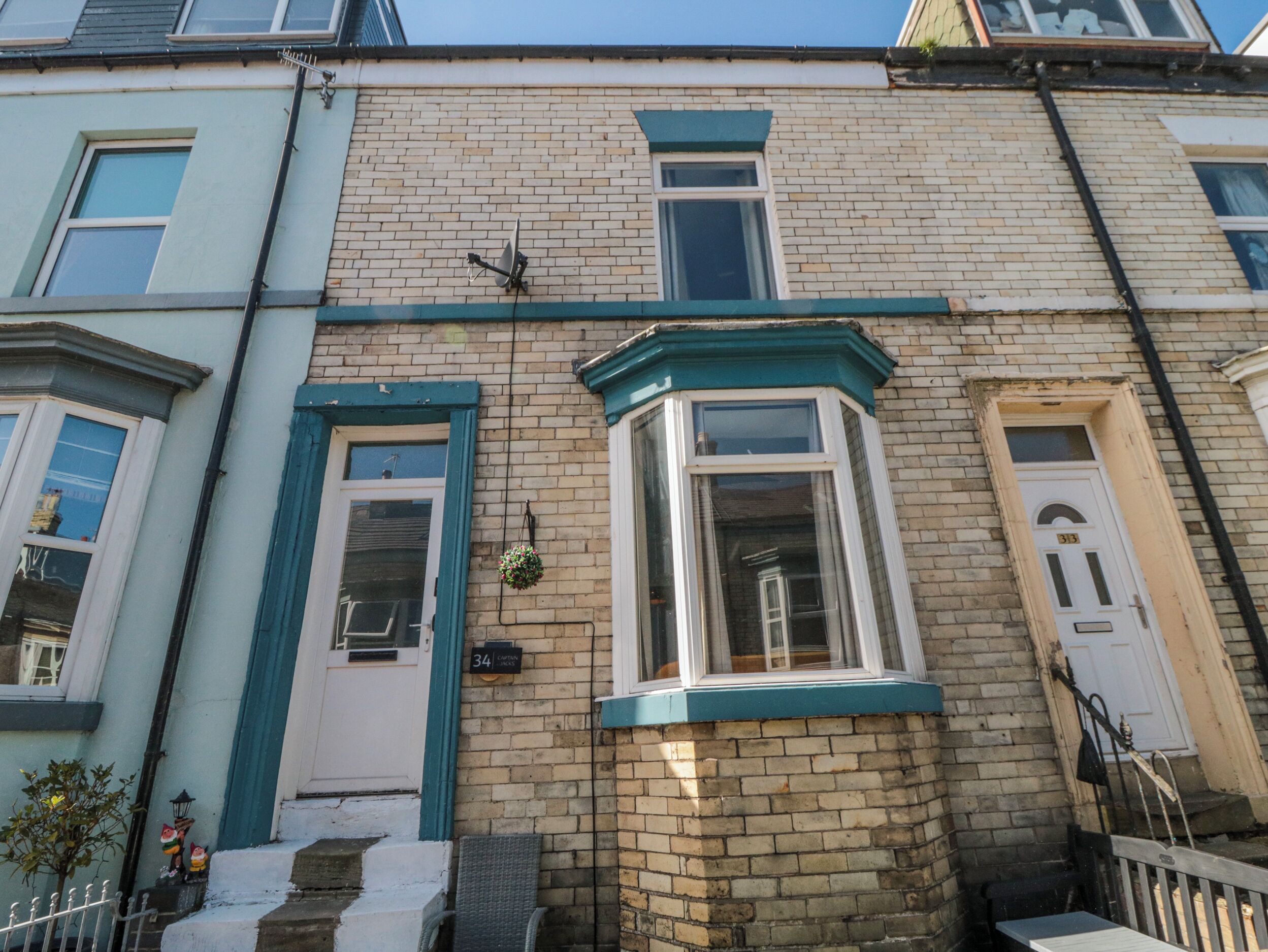 Une maison de ville d’angle avec une baie vitrée, une porte avec une plaque de numéro et un petit pot de fleurs suspendu.