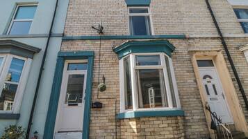 Une maison de ville d’angle avec une baie vitrée, une porte avec une plaque de numéro et un petit pot de fleurs suspendu.