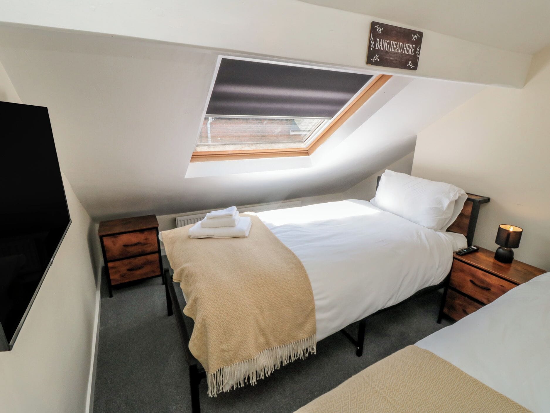 A bedroom with a skylight, a bed, a bedside table with a lamp, and a TV on the wall.