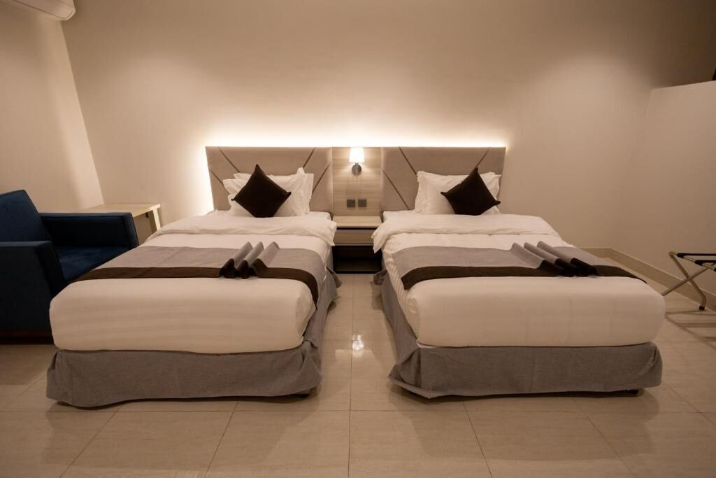 A hotel room with two beds, a bedside table, a chair, and a headboard with integrated lighting.