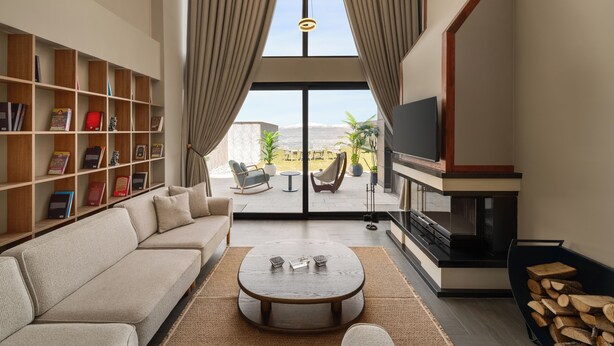 A modern living room with a sofa, coffee table, bookshelf, TV, fireplace, and a view through a glass door. - Ramada by Wyndham Erzincan Ergan (Erzincan)