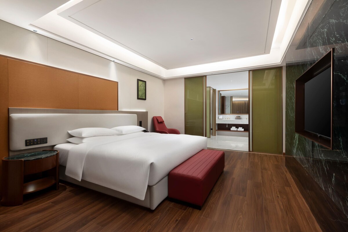 Una camera d'albergo moderna con un letto grande, una poltrona rossa, una TV e un bagno visibile tramite un separé in vetro.
