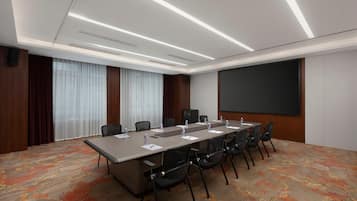 Un salón de conferencias con una larga mesa, sillas, una pantalla de proyector y un amplio ventanal con cortinas.