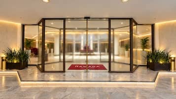 A hotel entrance with a glass door, a red mat with the hotel's name, and potted plants on either side.