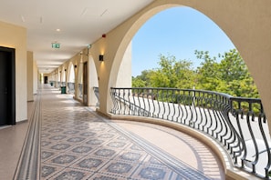 A corridor with patterned floor tiles, a curved balcony with a metal railing, and a clear view of greenery outside.
