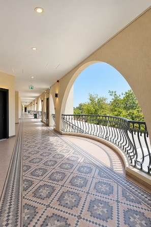 A corridor with patterned floor tiles, a curved railing, and a clear view of greenery outside.