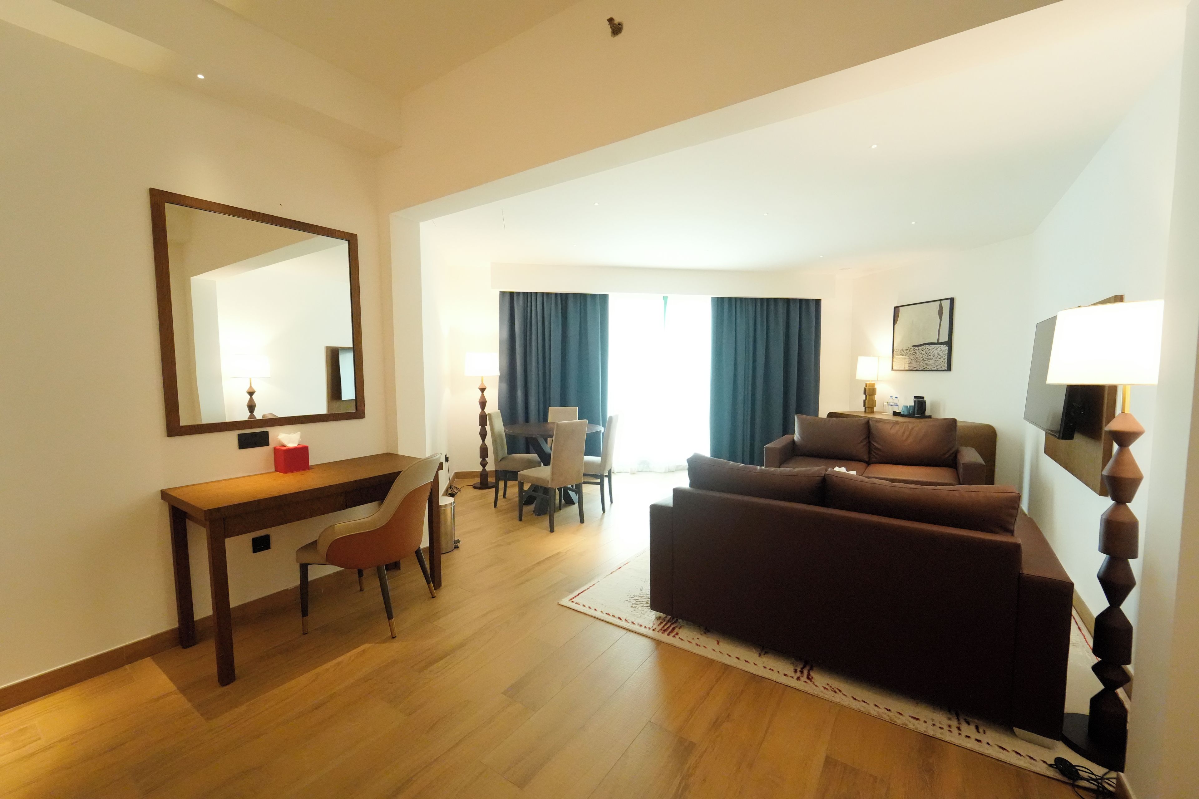 Quarto de hotel com uma escrivaninha de madeira, um sofá marrom, uma área de jantar com cadeiras e um espelho grande na parede.