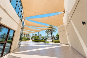 A covered walkway with a geometric floor pattern, leading to a building with large windows and a palm tree-lined path.