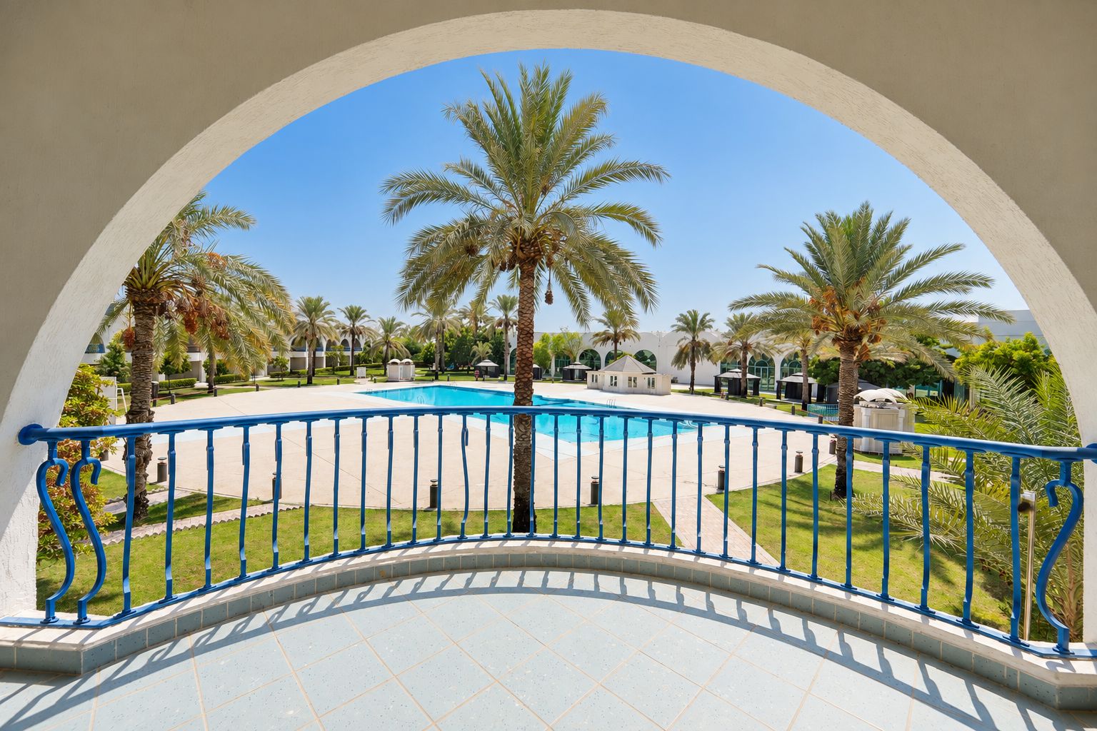A pool area with a clear blue sky, surrounded by palm trees and a white building with arches.