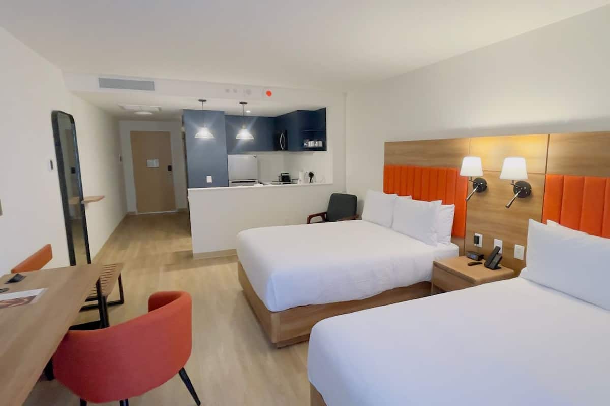 A hotel room with two beds, a desk, a chair, a kitchenette, and a bathroom.