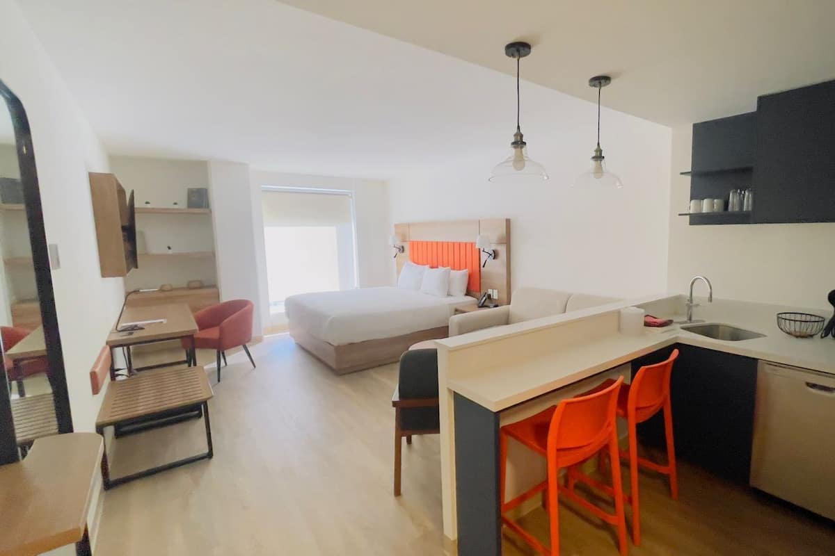 A modern hotel room with a kitchenette, a bed, a desk, and a dining area.