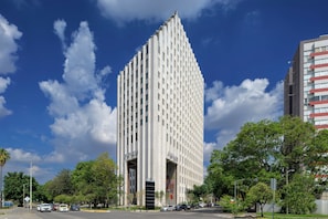 Un edificio alto y moderno con una fachada geométrica única, rodeado de vegetación y automóviles estacionados.