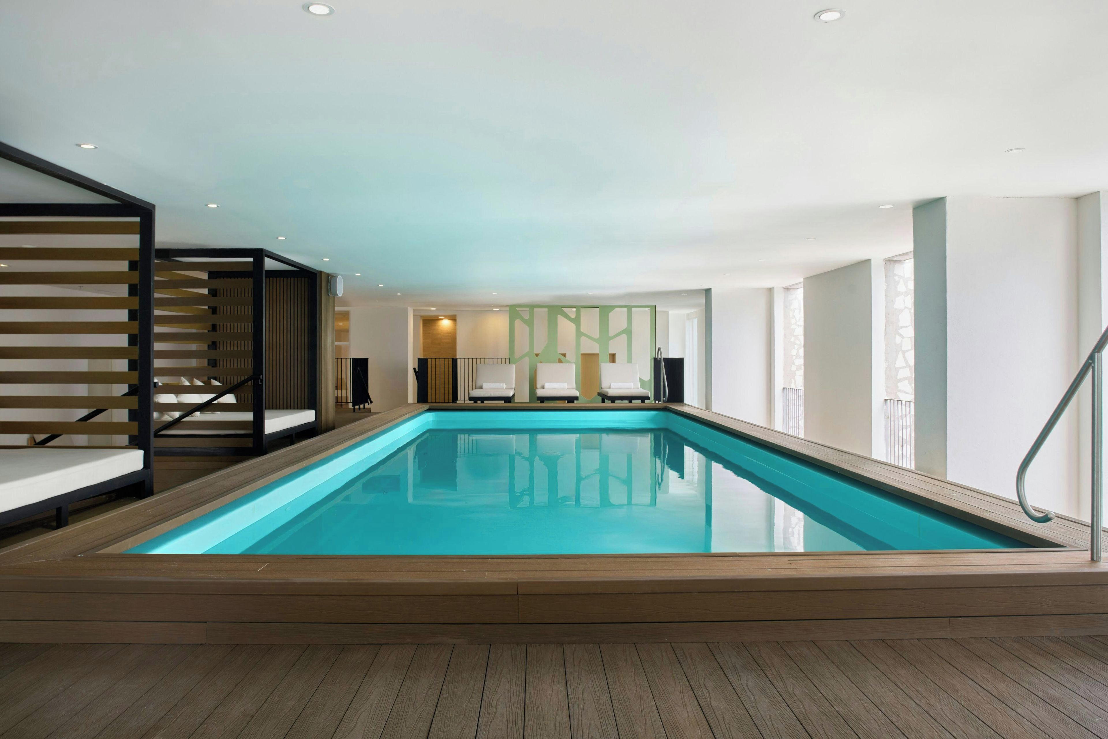 A modern indoor swimming pool with a wooden deck, a staircase with a black railing, and a view of the interior space.