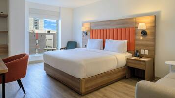 Quarto de hotel com uma cama grande, cabeceira de madeira, mesa de cabeceira com abajur, uma cadeira e vista para o exterior.
