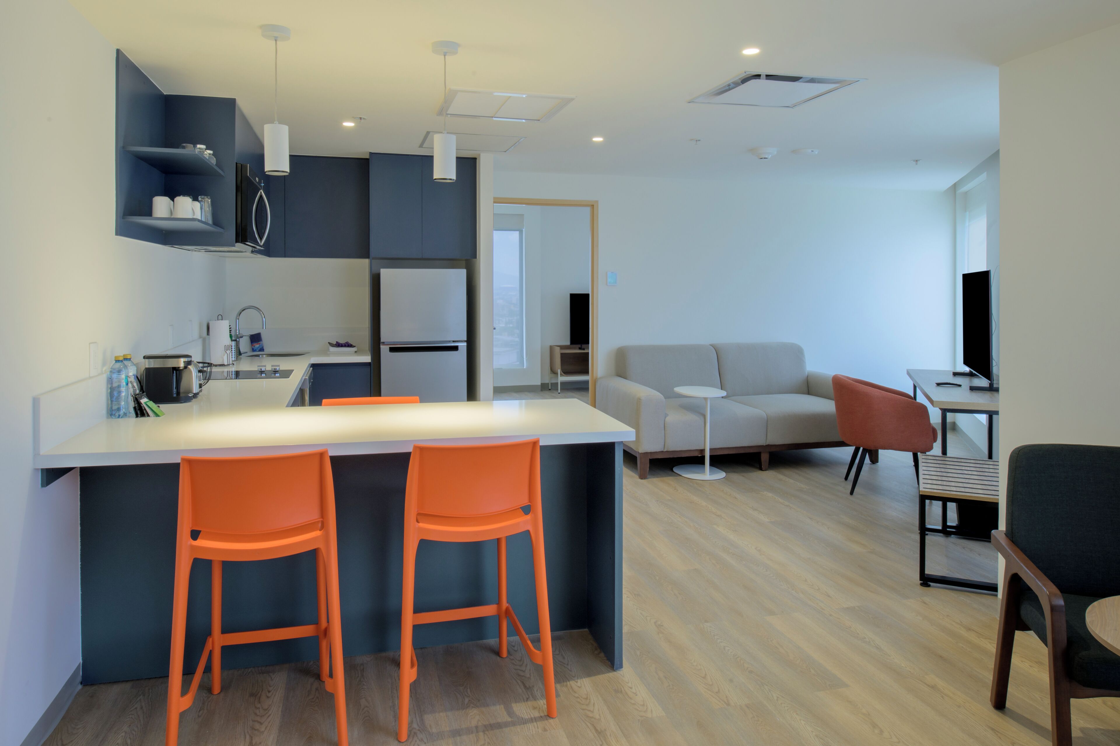 A modern kitchen with a central island, two orange bar stools, a grey sofa, and a television in a living area.