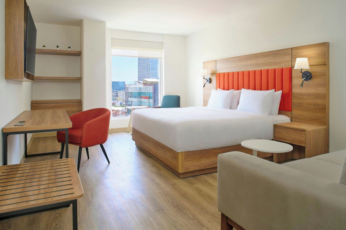 Une chambre d’hôtel avec un lit, un bureau, une chaise, une petite table et une vue sur la ville.