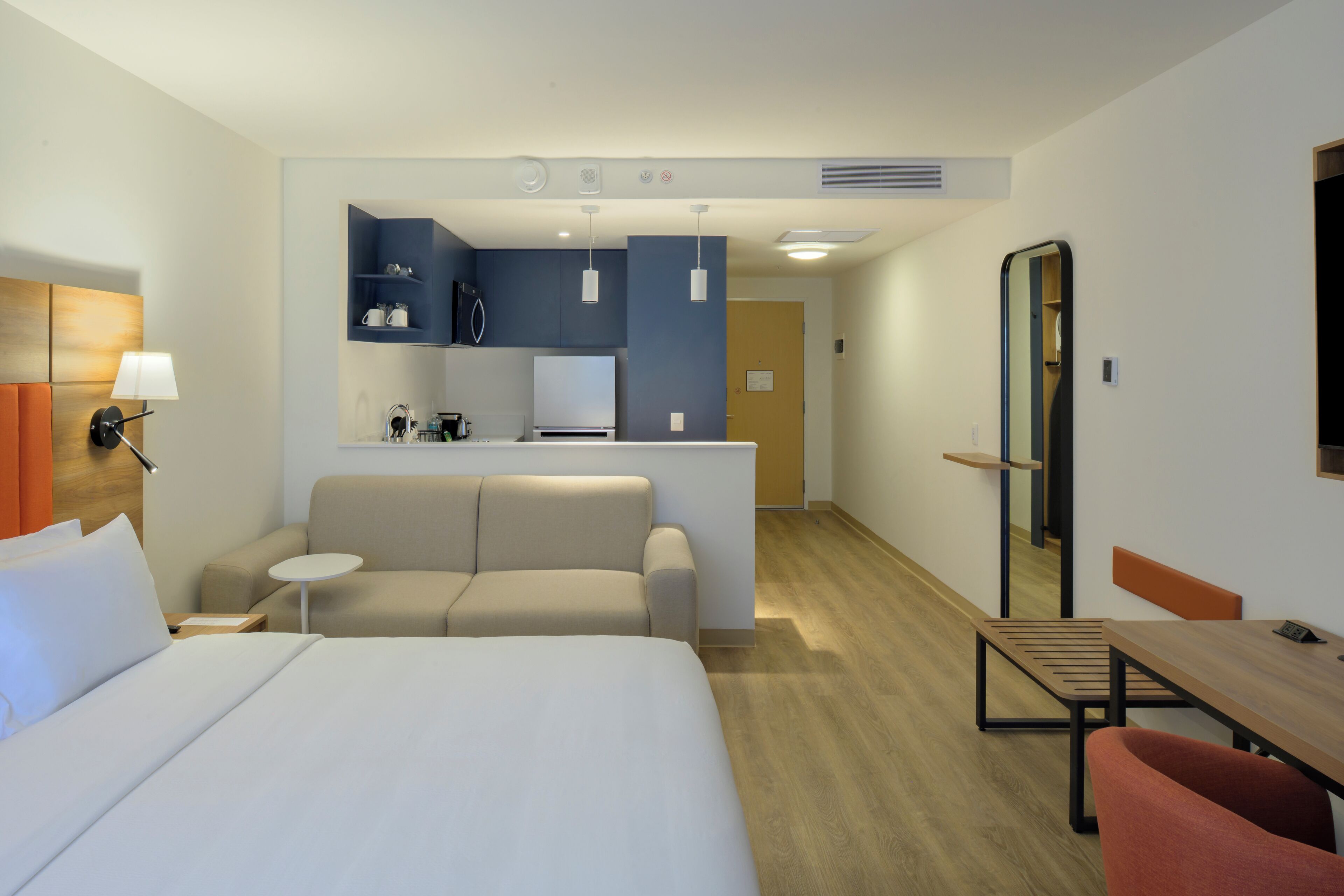A hotel room with a bed, a sofa, a small table, a desk, a mirror, and a kitchenette area.
