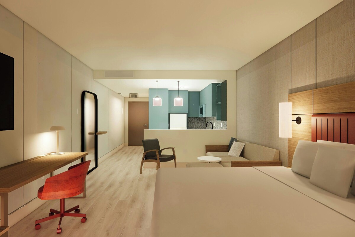 A modern hotel room with a wooden desk, a red chair, a bed, a small table, and a kitchenette in the background.