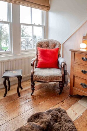 Ein Zimmer mit einem Holzsessel, einem roten Kissen, einem kleinen Holztisch, einer Holzkommode und Blick durch ein Fenster.