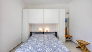 Un dormitorio con cama, armario blanco, lámparas de noche y una silla.