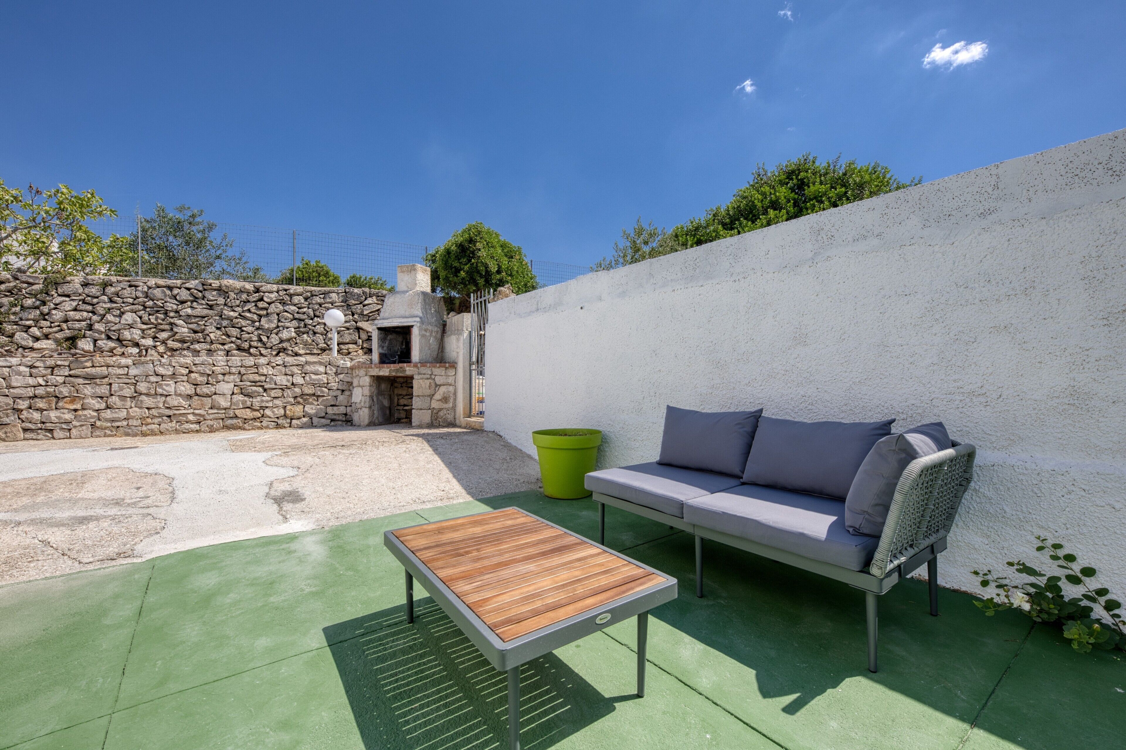 Une terrasse avec un mur en pierre, une table en bois, un canapé gris et une poubelle verte.