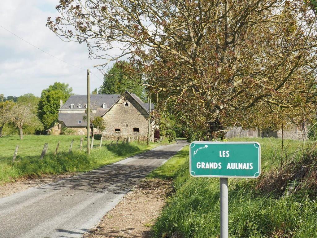 A rural road with a sign for 'LES GRANDS AULAIS' and a stone house in the background.