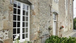A stone building with a large window, a door, and a small garden area with plants. - Les Grands-Aulnais (Astillé)