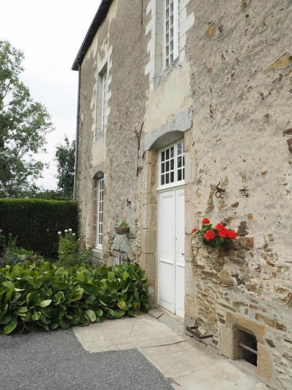 A stone building with a white door, a small window, and a flower box with red flowers. - Les Grands-Aulnais (Astillé)