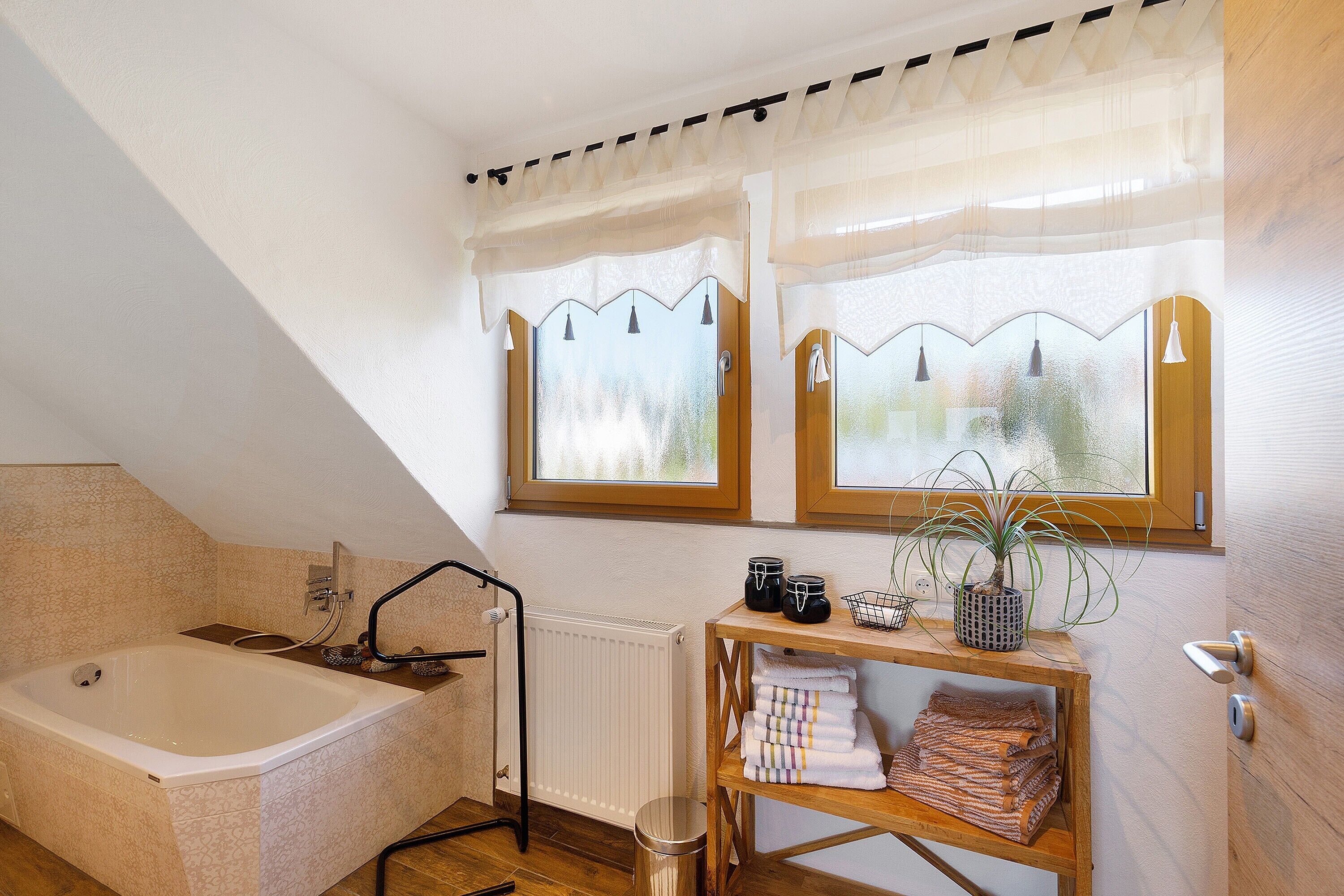 Une salle de bain avec une baignoire, un radiateur, une fenêtre avec des rideaux, une étagère avec des serviettes et une plante en pot.