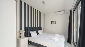 Un dormitorio moderno con pared a rayas, una cama con ropa de cama blanca, una mesita de noche, una lámpara y una ventana con cortinas.