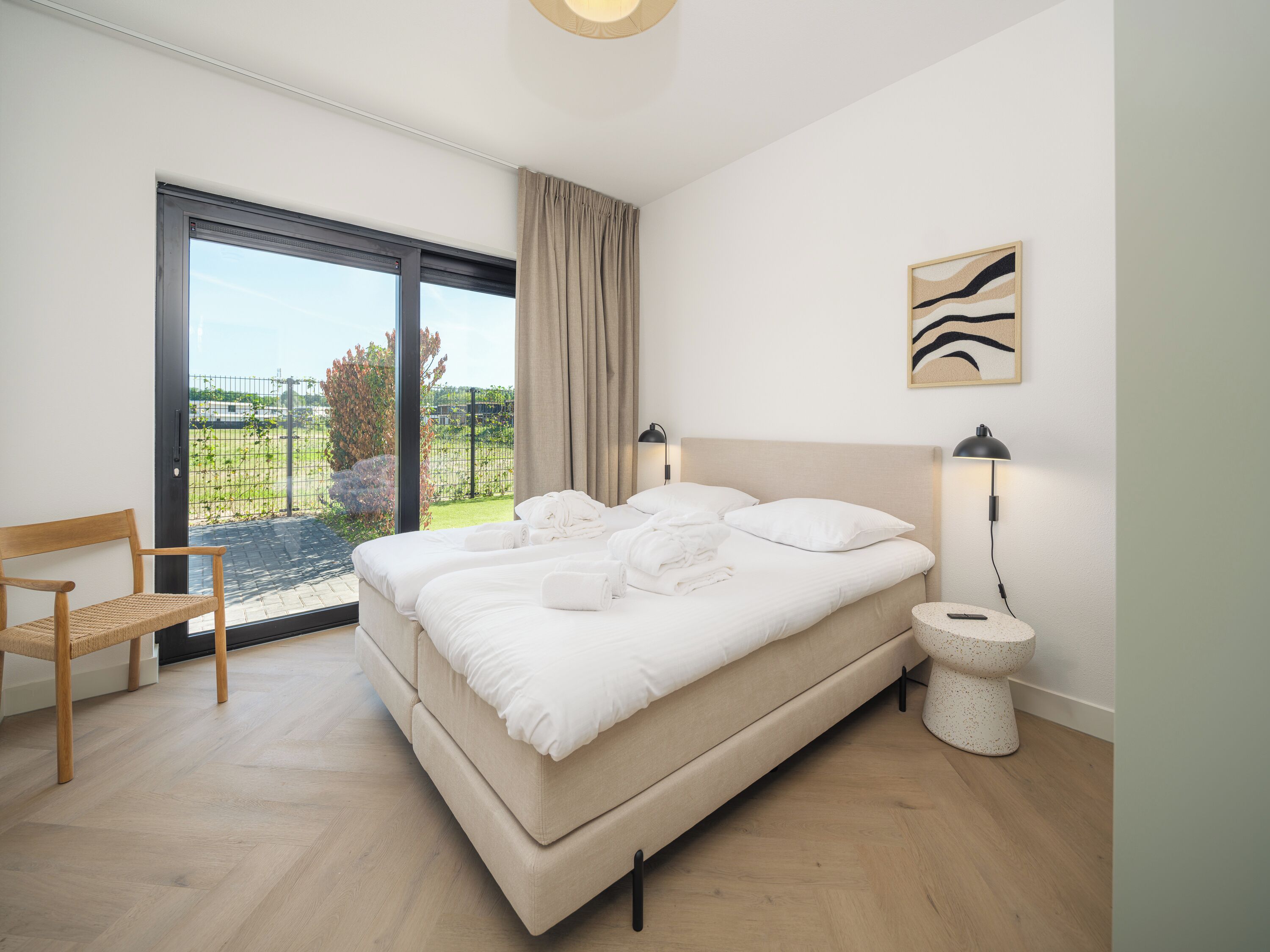 Une chambre avec un grand lit, une chaise en bois, une table de chevet et une vue sur un jardin, accessible par une porte coulissante.