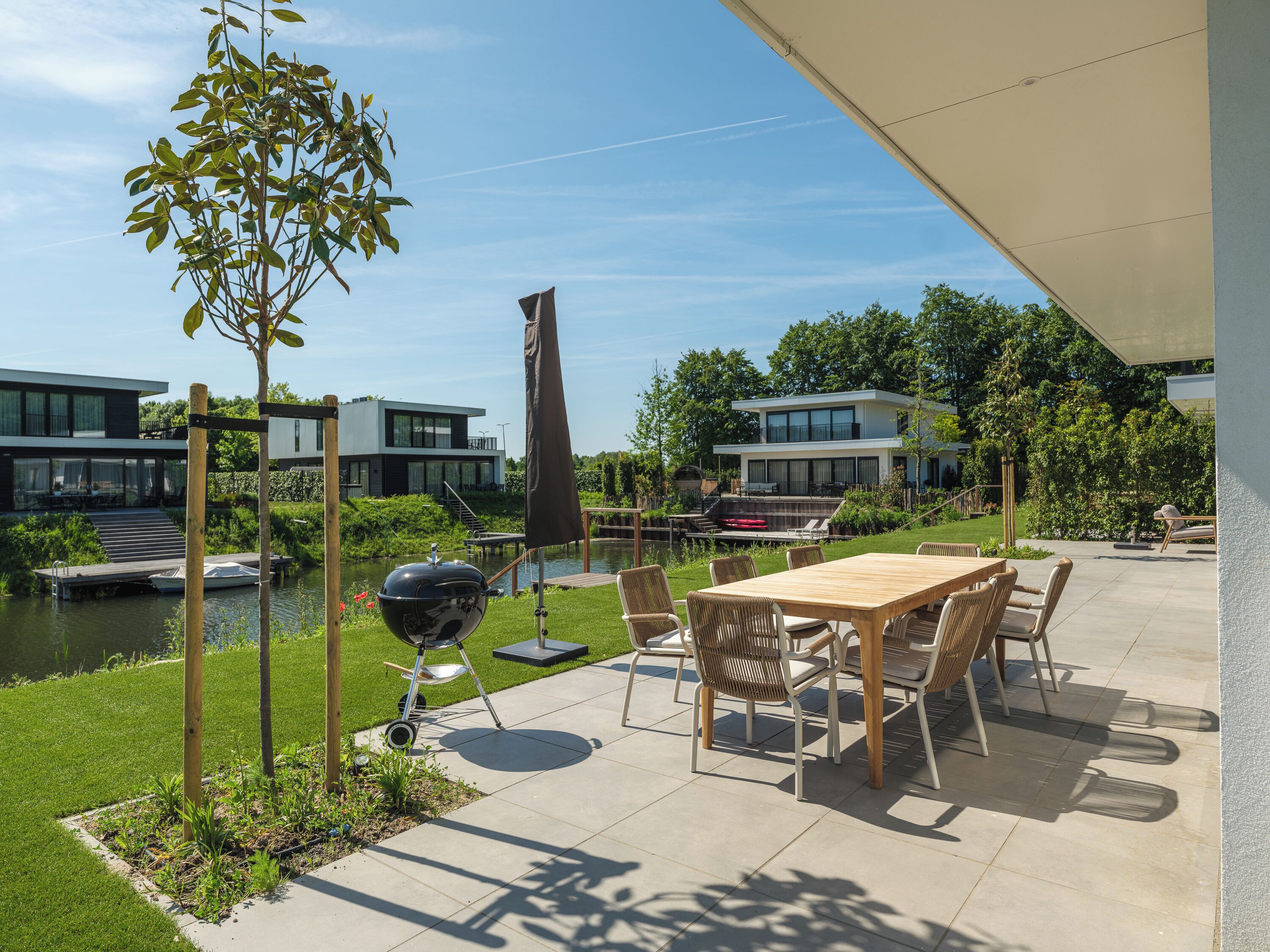 En terrasse med et bord og stole, en grill, en lille båd og et vandareal.