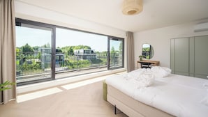 Et moderne soveværelse med en stor seng, et skrivebord, et spejl og udsigt til en balkon med grønne planter og bygninger.